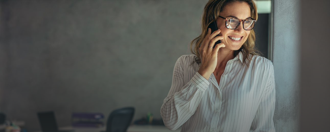 A person wearing glasses and a striped shirt is smiling while talking on a smartphone, standing indoors with a blurred office background.