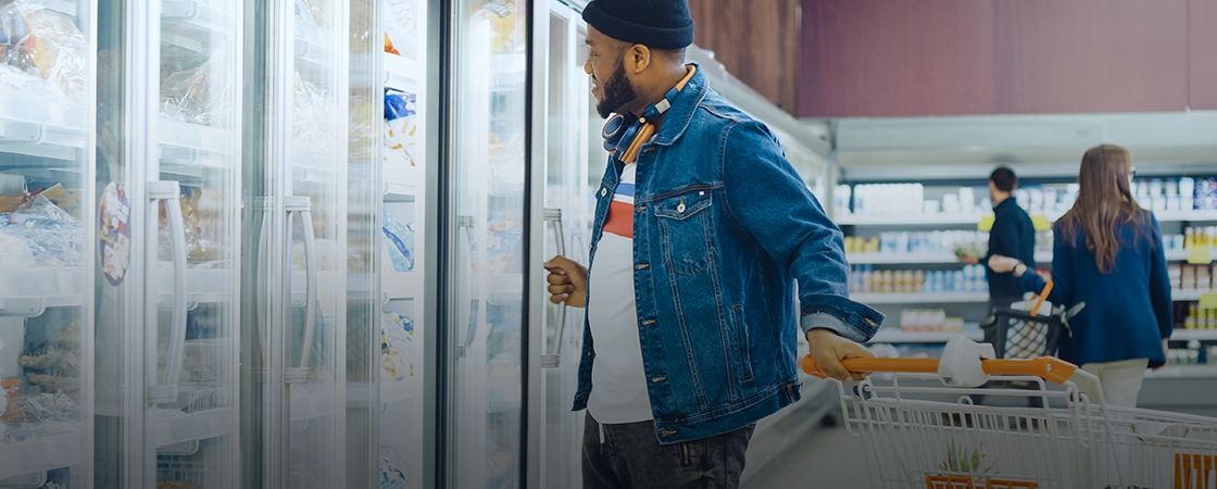 A person wearing a denim jacket and beanie is shopping in a supermarket. They are pushing a cart and opening a freezer door while looking inside. Shelves of groceries are visible in the background with another shopper nearby.