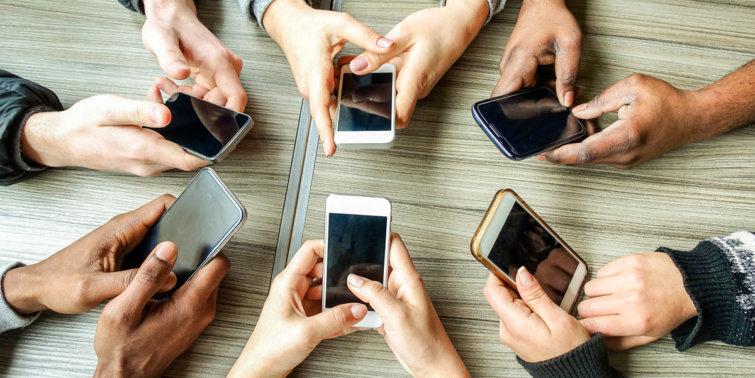 A group of people sitting around a table, each holding and looking at a smartphone. The phones are in various positions, and the table surface is wooden. The hands belong to different individuals, showing a diverse range of skin tones.