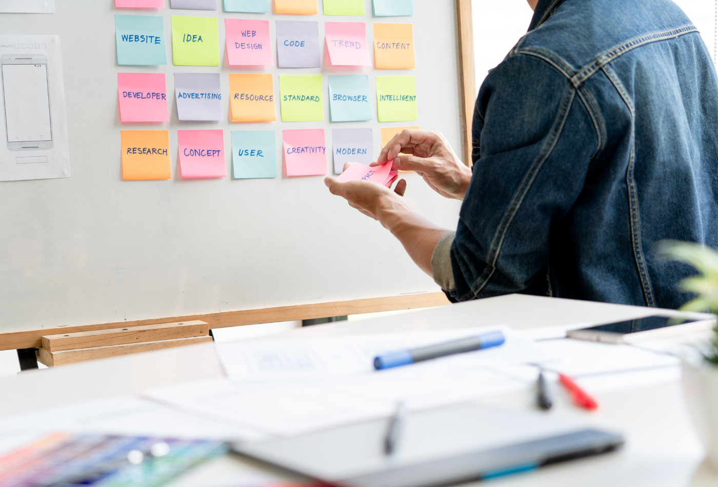 A person in a denim jacket places a sticky note on a whiteboard covered with colorful sticky notes containing various words. A desk in the foreground has papers, a smartphone, and pens.
