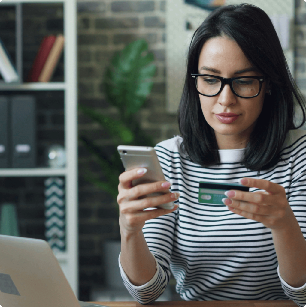 A woman in glasses and a striped shirt sits at a desk, holding a smartphone in one hand and a credit card in the other. A laptop is open in front of her, and a brick wall with shelves is visible in the background.