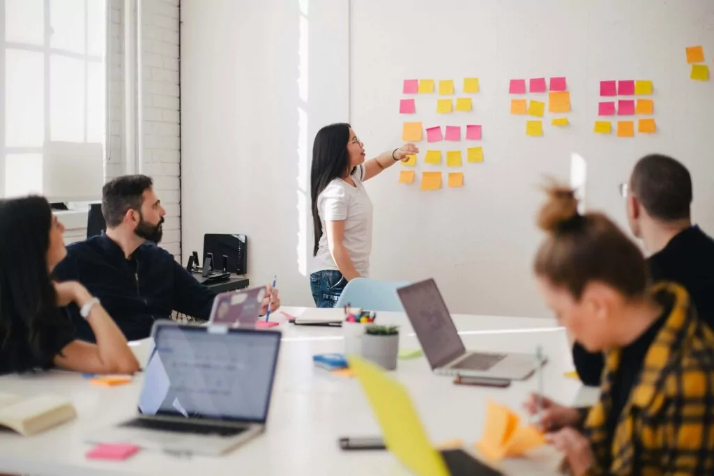 In a vibrant meeting room, focused professionals from the software industry attentively watch a woman point at a whiteboard adorned with colorful sticky notes, enhancing employee experience as they engage with laptops and notebooks at the table.