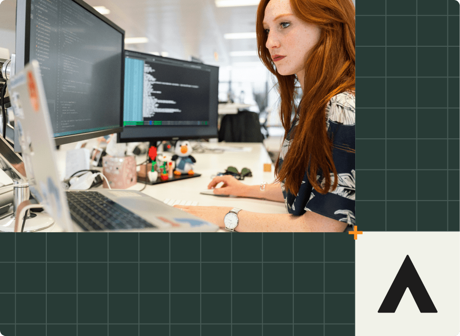 A woman with long red hair types on a keyboard at a desk with multiple monitors displaying code, focusing on software feedback. Office decor, a laptop, and a geometric design on the right side enhance the modern office setting.